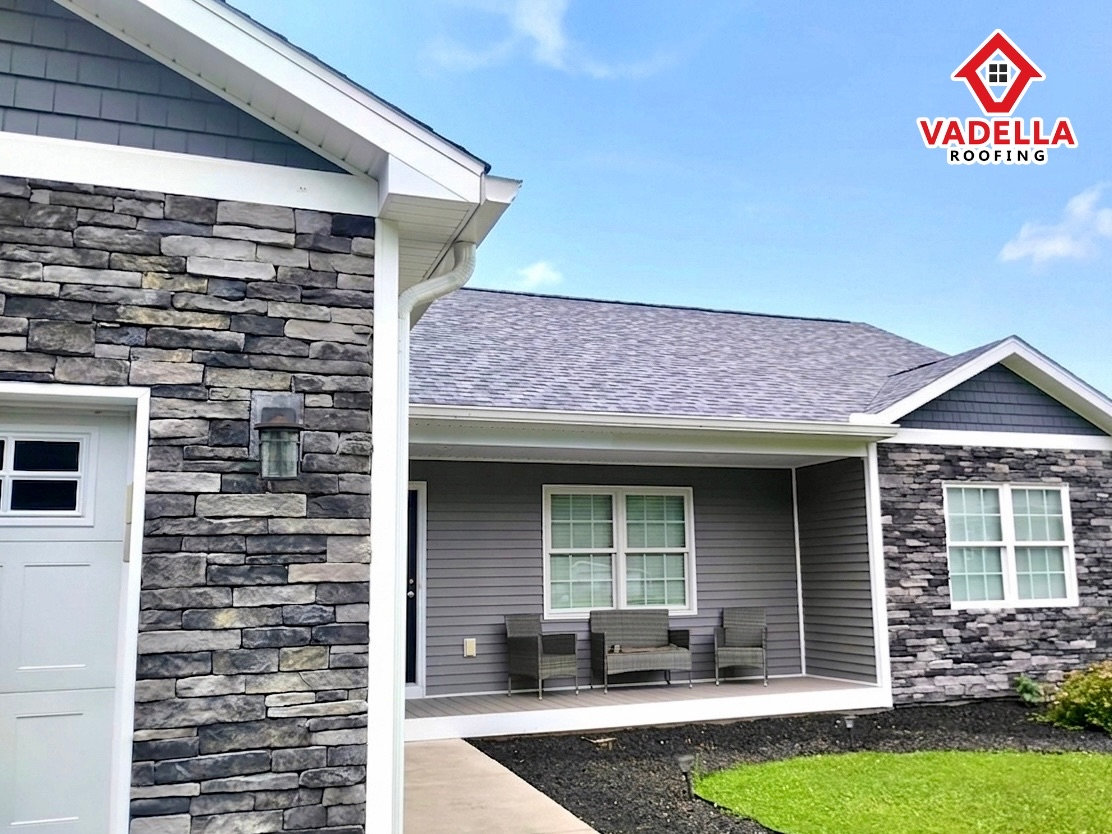 An exterior view of a modern ranch-style home featuring a new grey shingled roof and dark grey horizontal siding. The house is accented with stacked stone veneer in shades of grey and charcoal on the garage and front pillars. A covered porch area includes a grey outdoor seating set. In the top right corner, there is a logo for "VADELLA ROOFING."